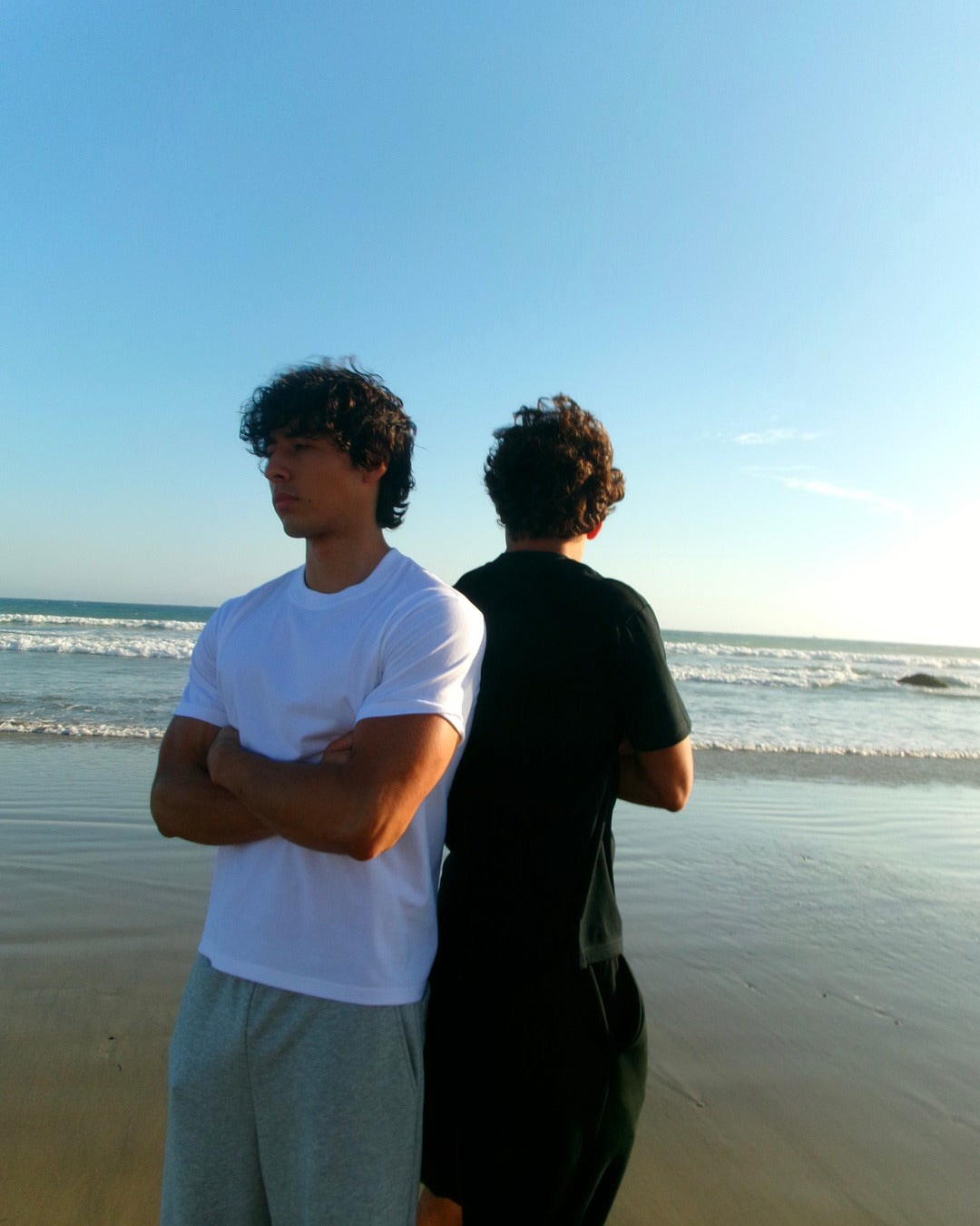 Two models in black and white SJ Goods shirts standing back to back near the ocean